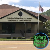 Exterior of the Yoknapatawpha County Sheriff's Department building with the Oxford Weekly Planet logo in the foreground