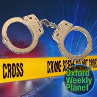 Handcuffs with crime scene tape and police lights in the background with the Oxford Weekly Planet in the foreground